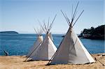 Three teepees (aka tipis) are lined up in a row in front of water. These teepees are used at a summer camp. Teepees were one form of traditional shelter used by native americans.