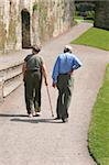 Elderly man and woman walking together along a path with the female holding a walking stick and the man holding his back as if in pain.