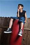 Young asian boy sitting on top of a punching bag outside beside a tall wooden fence resting his chin on hand wearing jeans and black tshirt