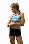 Attractive blond woman in great physical shape wearing black shorts and workout top standing with a measuring tape wrapped around waist looking at the measuring tape