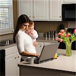 Caucasian woman holding baby  and typing on laptop computer in kitchen.