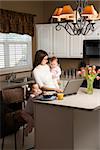 Caucasian mother holding baby  and typing on laptop computer with girl eating breakfast in kitchen.