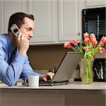 Caucasian man in suit using laptop computer and talking on cellphone in kitchen.