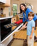 Hispanic mother and son putting cookies into oven and smiling at viewer.