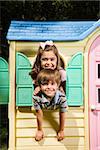 Hispanic boy and girl posing in window of playhouse and smiling at viewer.