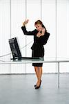 Caucasian businesswoman standing at computer desk waving on telephone.