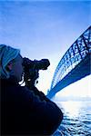 Caucasian teenage male photographing Sydney Harbour Bridge at dusk with view of Sydney Harbour, Australia.