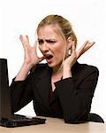 Attractive blond hair woman wearing business suit sitting in front of a computer with angry facial expression with hands up while looking at the computer as if it crashed or broke