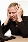 Attractive blond hair woman wearing business suit sitting in front of a computer with angry or confused facial expression with hands on face while looking at the computer as if it crashed or broke