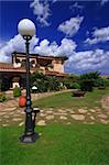 House/hotel in the beautiful summer garden with many plants and path - blue sky with clouds above and a lamp in the front