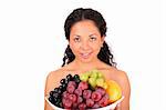 A smiling woman holds a plate with different kinds of fruits in her hands, standing on white background.