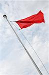A red flag flying from flag pole seen from below, clouds and blue sky beyond