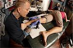 A tattoo artist applying his craft onto the lower leg of a pain-inflicted female.