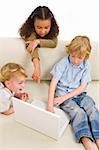 Three young children using a laptop computer while on a settee at home