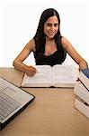 Young Hispanic woman sitting in front of desk with a pile of thick textbooks while reading one