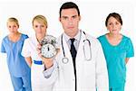 A male doctor backed by his medical team holding out an alarm clock ticking ever closer to 12 o'clock. The focus is on the clock face.