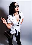 A high-key portrait about an attractive trendy lady with black hair who has a glamorous look. She is wearing sunglasses, black pants, a white coat and a stylish handbag.