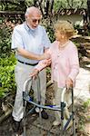 Senior couple outdoors.  She's in a walker and he's helping her.