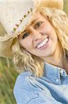 A beautiful blond haired blue eyed model wearing a straw cowboy hat sits amid tall grass illuminated by golden evening sunlight