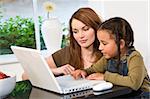 A beautiful young mother and her mixed race young daughter using a laptop computer at home in the kitchen.