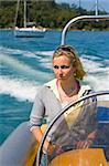 A stunningly beautiful young woman driving a powerboat away from her yacht at anchor off a Mediterranean coastline