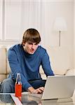 Brunette male using laptop while sitting on the coach wearing a long sleeved blue t-shirt, and drinking an orange soda. vertical