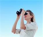 Blonde businesswoman looking through binoculars against blue sky