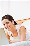 A beautiful young woman using a laptop in bed.  She is smiling at the camera. Vertically framed shot.