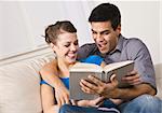 Attractive multi-ethnic couple reading and laughing together on their living room sofa. Horizontally framed shot.