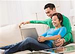 An attractive young couple on a couch, viewing a laptop screen. They are smiling. Horizontally framed shot.