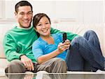 An attractive young couple sitting on a couch and watching television.  They are smiling directly at the camera.  The female is holding a remote.  Horizontally framed photo.