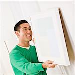 Asian male wearing a green sweater hanging art on wall. Square composition