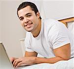 A man relaxing on a bed and using a laptop computer.  He has his fingers on the keyboard and is smiling at the camera. Square framed shot.