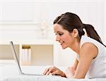 An attractive young woman lying on a bed and viewing a laptop screen.  She is smiling. Horizontally framed shot.