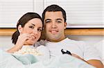 Couple sitting in bed together. The female is eating a bowl of popcorn and the male has a television remote. Horizontally framed photo.