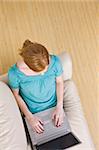 The above view of a young woman seated on a couch and working on a laptop. She has red hair.  Vertically framed shot.