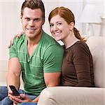 An attractive young couple sitting on a couch and smiling.  They are looking directly at the camera.  The male is holding a cell phone. Square composition.