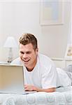 A young man lying on a bed and viewing a laptop screen.  He is laughing.  Vertically framed shot.