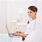 An attractive young female lying in bed and using a laptop.  She is smiling at the camera. Square framed photo.