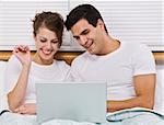 An attractive young couple lying in bed and viewing a laptop screen.  They are smiling. Horizontally framed photo.