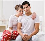 An attractive looking couple posing.  They are sitting on the edge of a bed and are wearing pajamas. They are smiling directly at the camera. Square framed photo.