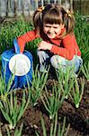 Little girl with watering can is sitting on the vegetable garden near onion patch