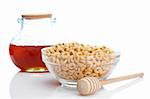 Honey jar, wooden drizzler and glass bowl of cheerios, reflected on white background