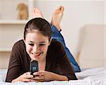 A young girl is laying on her bed and holding a cell phone.  She is smiling and looking away from the camera.  Square framed shot.