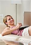 A young, smiling woman is laying on a couch and looking at a laptop.  Vertically framed shot.