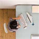 A businesswoman is sitting at a desk in an office.  She is working on a computer and looking at the screen.  Square framed shot.