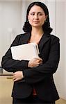 A businesswoman is standing in an office holding some paperwork.  She is looking at the camera.  Vertically framed shot.