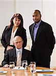 Drei Geschäftsleute für Foto posieren. Afroamerikaner Mann und Frau, stehend, männlich Senior vor ihnen sitzt. Vertikal.