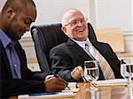 An elderly man and a young businessman are seated together at a desk in an office.  They are laughing and looking away from the camera.  Horizontally framed shot.