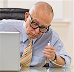 An elderly man is sitting at a desk in an office and is working on a laptop.  He is looking away from the camera.  Square framed shot.